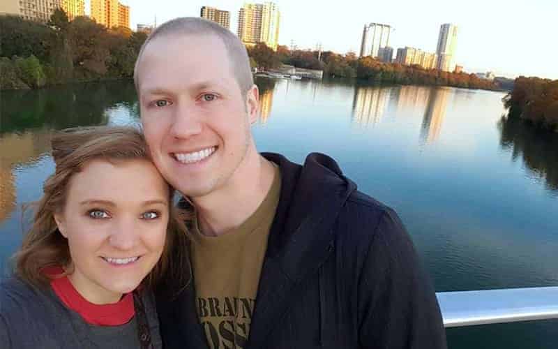 Smiling couple outdoors with city skyline and river in background.