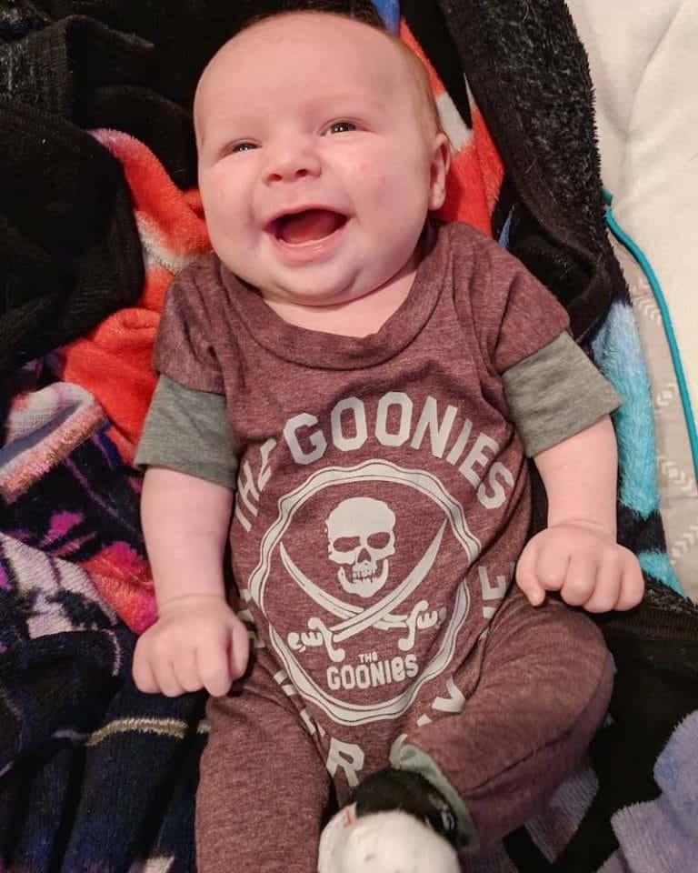 Say Hello To Rhonan 5 Happy baby wearing Goonies themed shirt, smiling and sitting in a cozy setting.